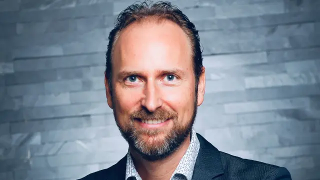 Philipp Günther steht vor einer grauen Wand und trägt ein gemustertes Hemd und ein Sakko. Er trägt einen Bart und lächelt in die Kamera.
