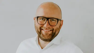 Rafael Schiwietz trägt ein weißes Hemd und eine Brille und lächelt vor einem weißen Hintergrund in die Kamera.