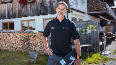 Reiner is standing in a Bosch polo shirt in front of a mountain hut with a wooden facade, holding a laptop in his hand.