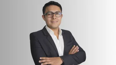 Rene is seen in front of a gray background and smiles into the camera. He is wearing glasses and a suit.