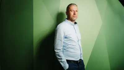 Porträt von Robert Ferencz, der vor einem grün-gemusterten Hintergrund steht. Er trägt ein Hemd, hat die Hände in die Hosentaschen gesteckt und blickt freundlich in Richtung Kamera.