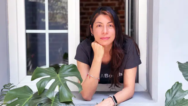 Selene Bermudez lehnt sich aus einem offenen Fenster mit weißem Rahmen nach draußen, hat ihren Kopf auf einen Arm gestützt und um sie herum sind Pflanzen sichtbar.