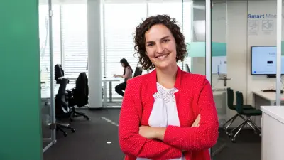 Theresa Best steht mit verschränkten Armen vor einem Büroraum. Sie trägt ein weißes Shirt und einen roten Blazer und lächelt in die Kamera.
