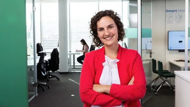 Theresa Best steht mit verschränkten Armen vor einem Büroraum. Sie trägt ein weißes Shirt und einen roten Blazer und lächelt in die Kamera.
