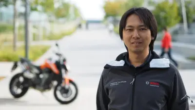 Yuki Oshida is wearing a Bosch jacket, standing outside and looking at the camera. The background is blurred, but trees and a motorcycle are still visible.