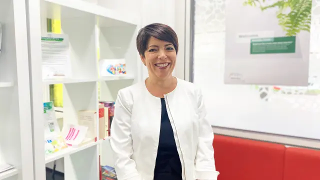 Yvonne Ongetta is wearing a white leather jacket over a dark blue shirt. She stands next to a white shelf and a red sofa and smiles into the camera.