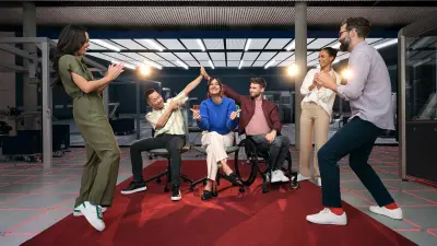 Sechs Personen sind auf einem roten Teppich in einem Labor zu sehen. Sie lachen alle und bejubeln sich gegenseitig.