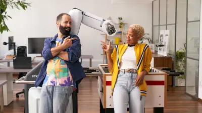 Um homem barbudo e uma mulher com cabelo loiro curto posam de forma divertida ao lado de um braço robótico num espaço de escritório moderno.