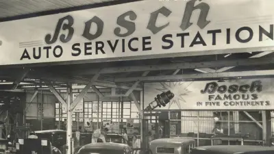 Car repair shop with many vehicles and mechanics, at the top of the picture large sign “Bosch Service Station” and below sign “Bosch famous in five Continents”
