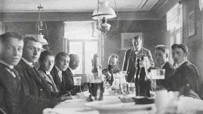 Nine men sit around a table set with dishes, glasses and drink bottles, one standing. All except the man with a full beard at the very back are looking at the photographer.