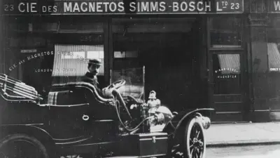 Historic open car in front of a building with a sign reading “Cie. des Magnetos Simms-Bosch”