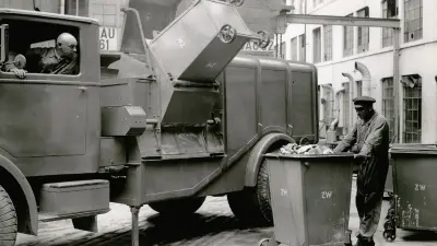 A garbage bin is being tipped into a vehicle. A man looks out the window. Another man stands next to the vehicle with a garbage bin.
