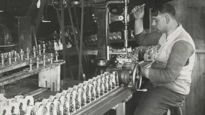 A man sits at a workbench with a lever in his hand. Next to him, in a row, are a dial and an ignition device.