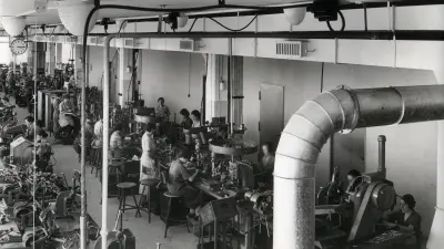 View from above of the ducts under the ceiling of a factory space. Under them sit women at workbenches.