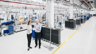 Zwei Personen in moderner Fertigungshalle, umgeben von Maschinen und automatisierten Arbeitsstationen.