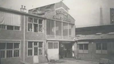 Factory building labeled Magnétos Simms-Bosch, with the top of the Eiffel Tower in the background.