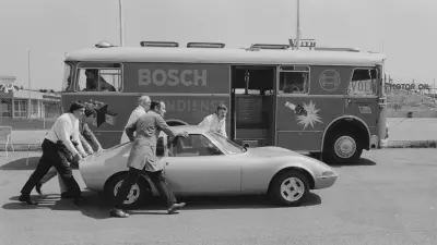 Several people push a vehicle on a race track. In the background, the service bus of the Bosch racing service.