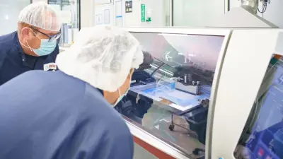 Zwei Menschen mit Schutzkleidung vor Produktfertigung hinter einer Glasscheibe
