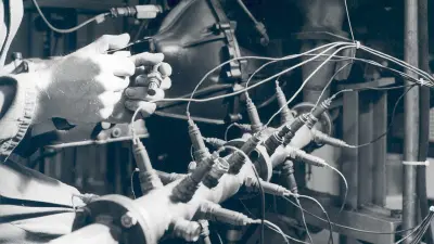 A tester’s hands can be seen holding a magnifying glass and a lambda sensor at a test apparatus for several lambda sensors in a pipe