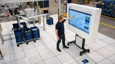 Anmutung einer Fabrikhalle mit gerastertem Boden. Ein Mann steht vor einem großen Bildschirm und deutet auf die dort angezeigte Grafik. Links von ihm befinden sich mehrere Kunststoffboxen, darüber ist ein Roboterarm, der die Inhalte der Boxen sortiert.