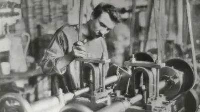 Ein Arbeiter steht in einer Werkhalle hinter einer Maschine und schaut prüfend auf das Werkstück, das zur Bearbeitung in die Maschine eingelegt ist.