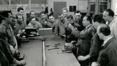 A group of men are standing in a workshop around a workbench. They are looking at a workpiece, which is held aloft by the master craftsman.