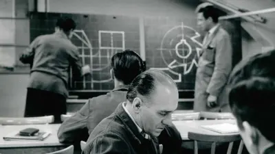 Two men are standing at a blackboard; one is drawing, and the other is watching. In front of them there are three men sitting at desks who are also looking at the blackboard or drawing themselves.