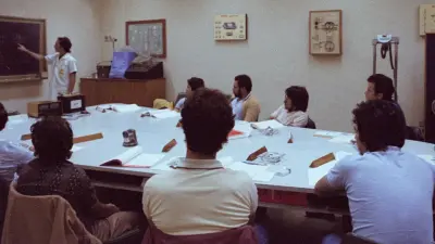 A number of men are sitting around a large white table and watching a man in a white apron explaining something at the blackboard.