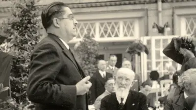 Robert Bosch mit anderen Personen am Tisch sitzend, schaut zu Redner, der links von ihm steht.