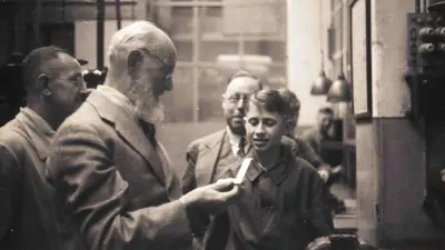 Robert Bosch inspects a workpiece made by an apprentice. The young apprentice and two other men look on.