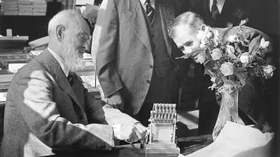 Robert Bosch sits at a table and demonstrates a small model of an injection pump. A man leans forward and watches.