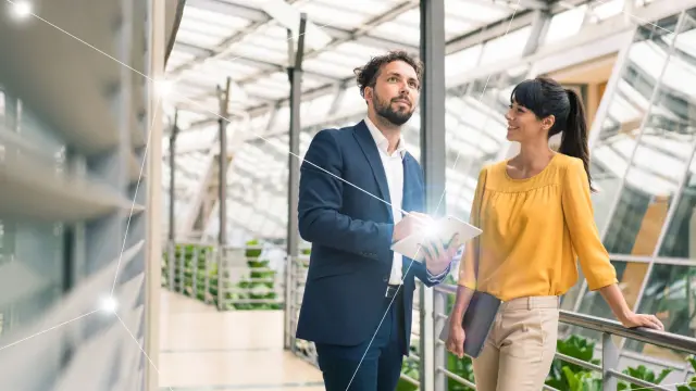 A man and a woman standing in a modern, glass-walled office building. The man is holding a tablet. There is a digital line around the tablet, adding a tech-inspired effect to the image.