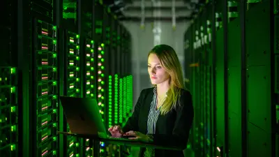 Female researcher working on a laptop in a modern data center with green LED lighting and server racks, representing Bosch's hybrid modeling and Industrial AI research.