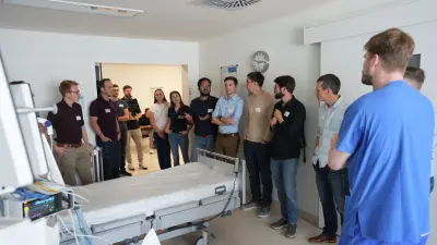 A group of about ten people stands in a medical examination room at the Bosch Health Campus, and observes a hospital bed with medical equipment.