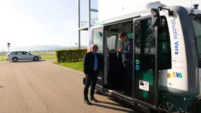 Two men standing next to the open entrance of an automated Bosch Research shuttle bus "Shuttle Vera", with a white car in the background.