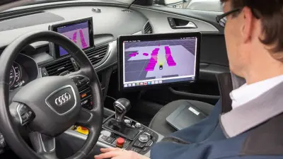 Im Innenraum eines Autos sitzt ein Forscher mit Brille auf der Fahrerseite und blickt auf einen Monitor, der sich rechts im Bild befindet. In der Mitte des Monitors ist ein abgebildetes Fahrzeug zu erkennen. Rechts und links von dem Fahrzeug sind violette Gebäudeblöcke zu sehen. Das Bild verdeutlicht die Bedeutung von Künstlicher Intelligenz (KI) bei der Umfelderkennung im Bereich des automatisierten Fahrens.