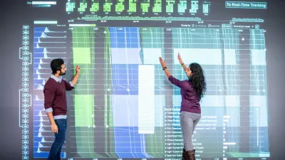 Eine Expertin und ein Experte von Bosch Research stehen vor einer PowerWall und analysieren Zahlen und Grafiken, die in verschiedenen Blau- und Grüntönen dargestellt sind. Die Expertin wendet sich dem Experten zu und streckt ihre Arme parallel nach oben, als würde sie ihm etwas erklären. Der Experte betrachtet nachdenklich die PowerWall und streckt ebenfalls seinen Arm nach oben.