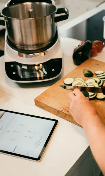 Bosch Kochassistenten in der Küche. Auf einem Holzbrett wird Gemüse geschnitten. Neben dem Kochassistenten liegt ein Tablet.