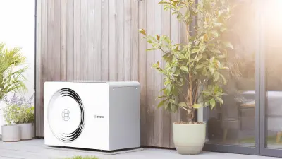 Das Foto steht für die Forschung an der Elektrifizierung von Gebäuden bei Bosch Research. Auf dem Bild ist ein modernes Haus zu sehen mit einer Bosch-Wärmepumpe links neben der Eingangstür, die das Bosch-Logo sichtbar trägt. Der Rasen, links im Bild, ist gepflegt und durch das Fenster des Hauses ist eine moderne Küche erkennbar.