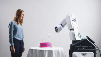 Eine Bosch Mitarbeiterin und der APAS-Assistenten feiern gemeinsam „10 Jahre Industrie 4.0“ mit einem Kuchen inklusive brennender Wunderkerze. 