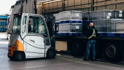 A man in a forklift loads shrink-wrapped pallets of goods onto the trailer of a semi-truck. A second man wearing a reflective vest stands beside the truck.
