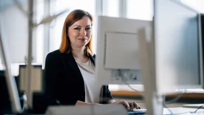 Juliane Lüder, a woman with red hair wearing a blazer, sits at a desk in a modern office, smiling at the camera. Two computer monitors are in front of her.