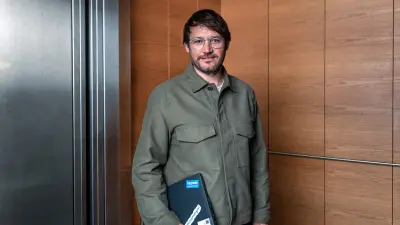 A man stands in an elevator, holding a laptop under his arm.