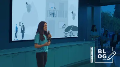 Shabnam Ghaffarzadegan in front of a monitor presenting Intelligent Audio Analytics. She is wearing a blue shirt and black jeans with black boots. The camera is facing her diagonally. She is holding a microphone in her left hand and is gesturing with her right hand. She is standing confidently as she is talking to the non-visible audience. On the wall behind her you can see a huge monitor with a slide about Human Machine Collaboration. Underneath the monitor you can see a row with cabinets. In the background you can see other team members standing and sitting while listening to Shabnam.