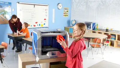 Eine Schülerin steht in einem Klassenzimmer vor einem 3D-Drucker und betrachtet lächelnd einen ausgedruckten Gegenstand