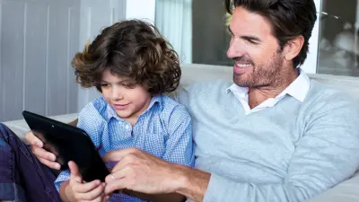 Sitting on a couch, a father uses a tablet to explain something to his son