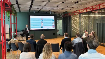 Speaker presents slides to a seated audience in a modern conference space with wood floors and green walls.