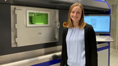 Eine Frau mit rötlichen, mittellangen Haaren steht in der rechten Bildhälfte. Sie ist bis zur Hüfte zu sehen und lächelt in die Kamera. Sie steht vor einer großen Maschine mit der Aufschrift SLM 500. Rechts an der Maschine befindet sich ein Computer-Bildschirm, in der Mitte eine kleine Tür mit Sichtfenster und grüner Beleuchtung.