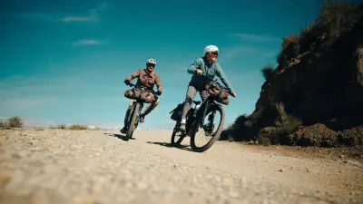 Two people ride loaded bicycles on a wide gravel road in hilly terrain.