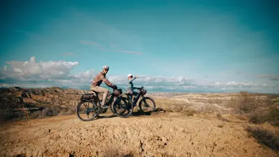 Two people with bicycles stand on a ridge overlooking a vast mountain landscape.
